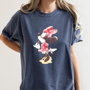 Puede incluir: Una camiseta azul oscuro con un gr&aacute;fico de Minnie Mouse con un lazo rojo y sosteniendo un l&aacute;piz labial. El gr&aacute;fico es de estilo vintage.