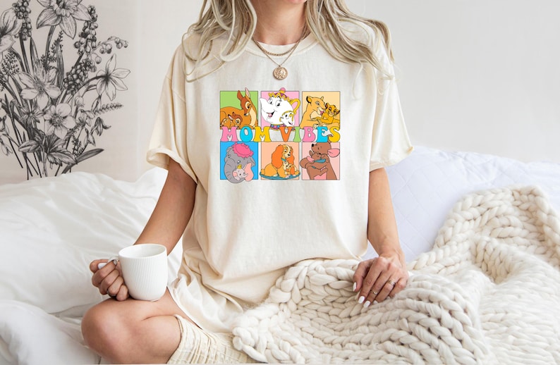 May include: A white t-shirt with a graphic print of Disney characters in colorful squares. The text "MOM VIBES" is printed in a colorful font across the squares.