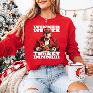 May include: Red Christmas sweater with a white graphic of a person holding a turkey dinner. The text on the sweater reads "Winner Winner Turkey Dinner".