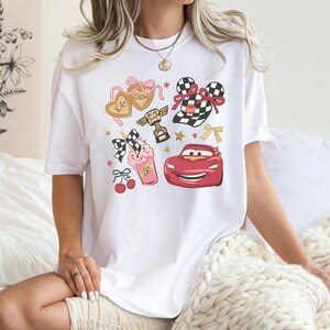 May include: White t-shirt featuring a colorful graphic design with cartoon elements. The design includes a red race car, a checkered flag, a milkshake, and heart-shaped cookies. The shirt has a crew neck and short sleeves.