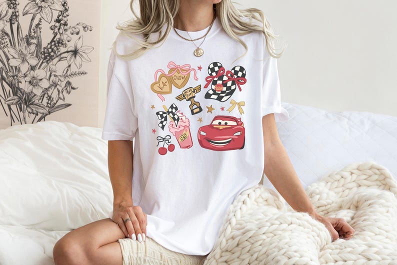 May include: White t-shirt featuring a colorful graphic design with cartoon elements. The design includes a red race car, a checkered flag, a milkshake, and heart-shaped cookies. The shirt has a crew neck and short sleeves.