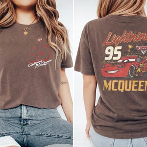 May include: A brown short-sleeve t-shirt featuring a small Lightning McQueen graphic on the front left chest and a large graphic on the back displaying a red race car, the number 95, a trophy, the Cars logo, and the text Lightning McQueen.