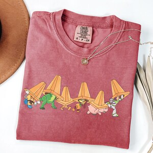 Puede incluir: Una camiseta Comfort Colors granate con un gr&aacute;fico de personajes de dibujos animados que llevan conos de tr&aacute;fico naranjas. Un collar dorado con un colgante est&aacute; en la camisa. Un sombrero marr&oacute;n y una planta seca est&aacute;n en el fondo.