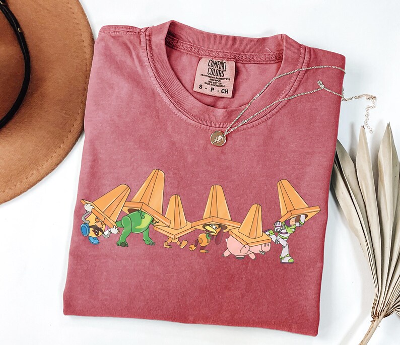 Puede incluir: Una camiseta Comfort Colors granate con un gr&aacute;fico de personajes de dibujos animados que llevan conos de tr&aacute;fico naranjas. Un collar dorado con un colgante est&aacute; en la camisa. Un sombrero marr&oacute;n y una planta seca est&aacute;n en el fondo.