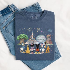 May include: A navy blue t-shirt featuring a colorful illustration of Disney characters and landmarks, including Cinderella's castle, Spaceship Earth, and the Tower of Terror. The shirt is laid on top of a pair of blue jeans.