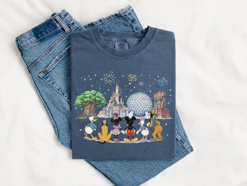 May include: A navy blue t-shirt featuring a colorful illustration of Disney characters and landmarks, including Cinderella's castle, Spaceship Earth, and the Tower of Terror. The shirt is laid on top of a pair of blue jeans.