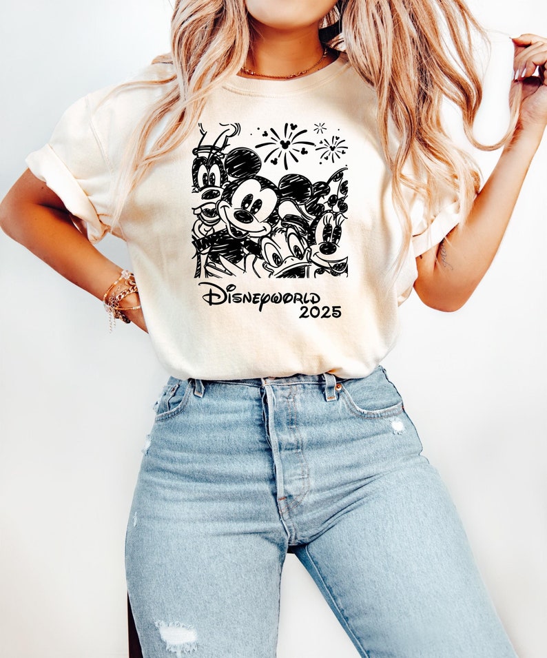 K&ouml;nnte beinhalten: Ein wei&szlig;es T-Shirt mit einer Schwarzwei&szlig;-Skizze von Mickey Mouse, Donald Duck und Goofy. Das Shirt tr&auml;gt au&szlig;erdem den Text "DISNEYWORLD 2025" und Feuerwerk.