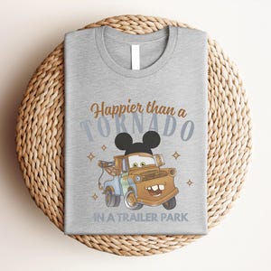 Puede incluir: Una camiseta gris jaspeado con el gr&aacute;fico de una gr&uacute;a marr&oacute;n con orejas de Mickey Mouse. El texto Happier than a TORNADO IN A TRAILER PARK aparece en letras marrones y azules, decorado con peque&ntilde;as formas de estrellas alrededor.