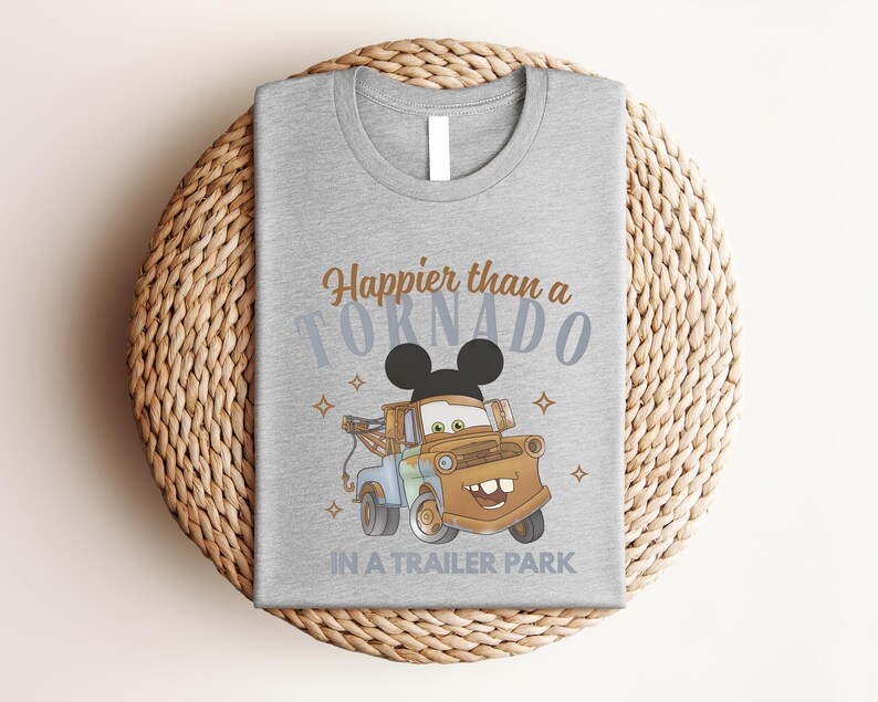 Puede incluir: Una camiseta gris jaspeado con el gr&aacute;fico de una gr&uacute;a marr&oacute;n con orejas de Mickey Mouse. El texto Happier than a TORNADO IN A TRAILER PARK aparece en letras marrones y azules, decorado con peque&ntilde;as formas de estrellas alrededor.