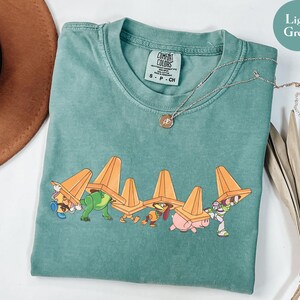 Puede incluir: Camiseta verde claro Comfort Colors con un gr&aacute;fico de dibujos animados de personajes de Toy Story que llevan conos de tr&aacute;fico naranjas. La camiseta tiene cuello redondo y mangas cortas. Un collar plateado con un peque&ntilde;o colgante est&aacute; drapeado sobre la camiseta.