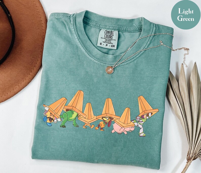 Puede incluir: Camiseta verde claro Comfort Colors con un gr&aacute;fico de dibujos animados de personajes de Toy Story que llevan conos de tr&aacute;fico naranjas. La camiseta tiene cuello redondo y mangas cortas. Un collar plateado con un peque&ntilde;o colgante est&aacute; drapeado sobre la camiseta.