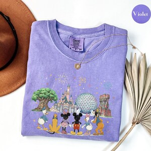 May include: A violet Comfort Colors t-shirt featuring a colorful graphic of Disney characters and landmarks. The design includes Mickey and Minnie Mouse, Donald and Daisy Duck, Goofy, and Pluto. The shirt has a crew neck and short sleeves.
