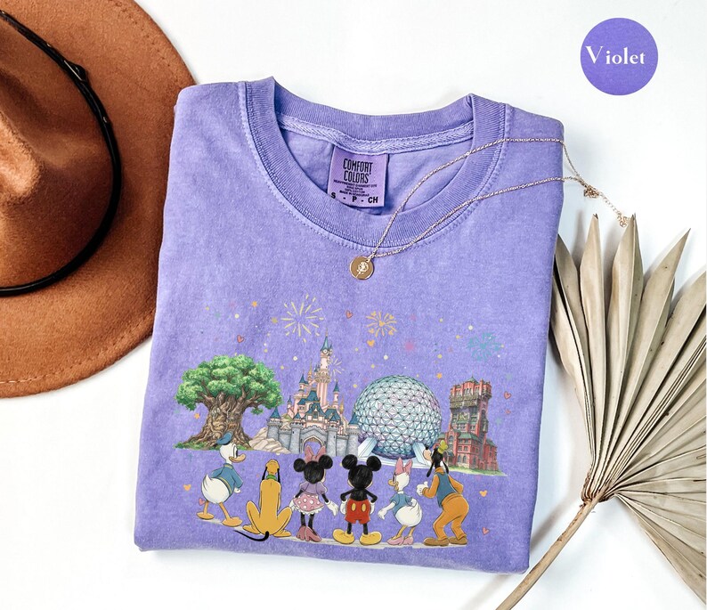 May include: A violet Comfort Colors t-shirt featuring a colorful graphic of Disney characters and landmarks. The design includes Mickey and Minnie Mouse, Donald and Daisy Duck, Goofy, and Pluto. The shirt has a crew neck and short sleeves.