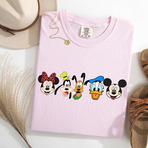 May include: Pink t-shirt with a graphic of Mickey Mouse, Minnie Mouse, Goofy, Pluto, and Donald Duck. The characters are all smiling and looking at the viewer.