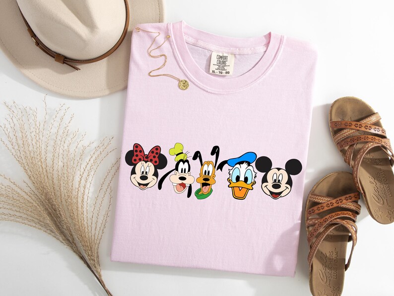 May include: Pink t-shirt with a graphic of Mickey Mouse, Minnie Mouse, Goofy, Pluto, and Donald Duck. The characters are all smiling and looking at the viewer.