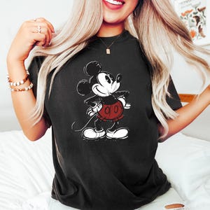 May include: A black t-shirt with a vintage-style illustration of Mickey Mouse in black and white. Mickey is wearing red shorts with white buttons.