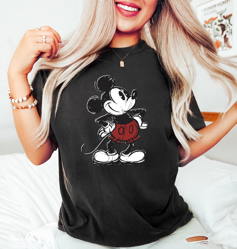 May include: A black t-shirt with a vintage-style illustration of Mickey Mouse in black and white. Mickey is wearing red shorts with white buttons.