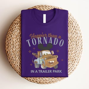 Puede incluir: Una camiseta morada con un gr&aacute;fico de una gr&uacute;a marr&oacute;n que lleva orejas de rat&oacute;n negras. El texto dice Happier than a TORNADO IN A TRAILER PARK en una mezcla de letras doradas y grises, acentuado con peque&ntilde;os iconos de estrellas.