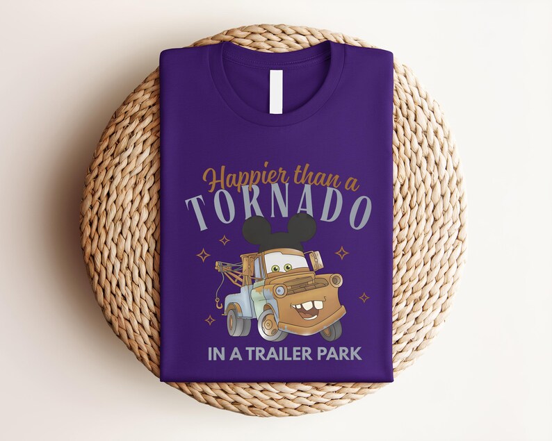 Puede incluir: Una camiseta morada con un gr&aacute;fico de una gr&uacute;a marr&oacute;n que lleva orejas de rat&oacute;n negras. El texto dice Happier than a TORNADO IN A TRAILER PARK en una mezcla de letras doradas y grises, acentuado con peque&ntilde;os iconos de estrellas.