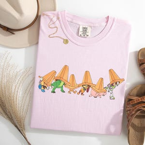 Puede incluir: Camiseta rosa claro con un dibujo animado de personajes con conos de tr&aacute;fico naranjas. La camiseta se combina con sandalias marrones, un sombrero beige y un collar dorado. La camiseta est&aacute; etiquetada XL-TG-EG.