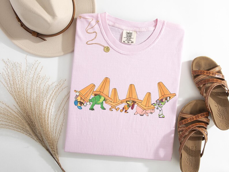 Puede incluir: Camiseta rosa claro con un dibujo animado de personajes con conos de tr&aacute;fico naranjas. La camiseta se combina con sandalias marrones, un sombrero beige y un collar dorado. La camiseta est&aacute; etiquetada XL-TG-EG.