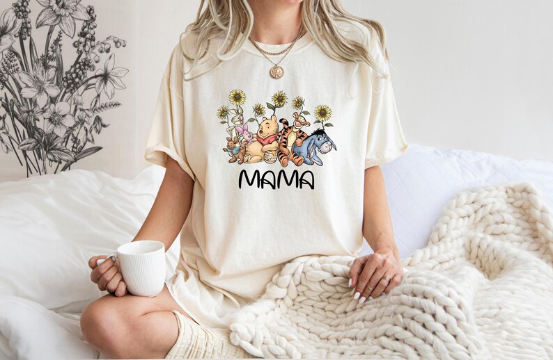 Puede incluir: Una camiseta blanca con un gr&aacute;fico de Winnie de Pooh y sus amigos, incluyendo Piglet, Tigger y Eeyore, rodeados de girasoles. El texto "MAMA" est&aacute; impreso debajo del gr&aacute;fico.