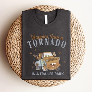 Puede incluir: Una camiseta gris oscuro con un gr&aacute;fico de una gr&uacute;a marr&oacute;n con orejas de rat&oacute;n. El texto dice Happier than a TORNADO IN A TRAILER PARK en letras naranjas, azul claro y blancas, decorado con peque&ntilde;as formas de estrellas de cuatro puntas.