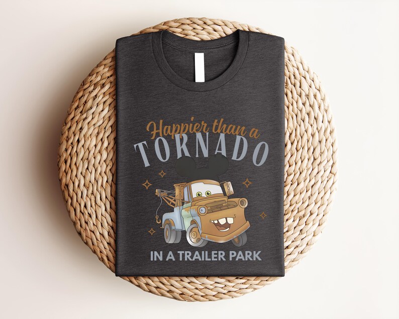 Puede incluir: Una camiseta gris oscuro con un gr&aacute;fico de una gr&uacute;a marr&oacute;n con orejas de rat&oacute;n. El texto dice Happier than a TORNADO IN A TRAILER PARK en letras naranjas, azul claro y blancas, decorado con peque&ntilde;as formas de estrellas de cuatro puntas.