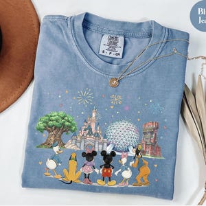 May include: Blue jean Comfort Colors t-shirt featuring a colorful graphic of Disney characters and landmarks. The design includes Mickey Mouse, Minnie Mouse, Donald Duck, and Goofy, with fireworks and iconic park structures. A gold necklace rests on the shirt.
