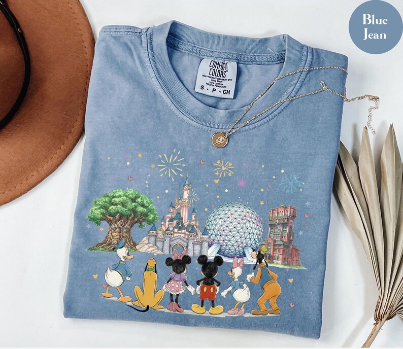 May include: Blue jean Comfort Colors t-shirt featuring a colorful graphic of Disney characters and landmarks. The design includes Mickey Mouse, Minnie Mouse, Donald Duck, and Goofy, with fireworks and iconic park structures. A gold necklace rests on the shirt.