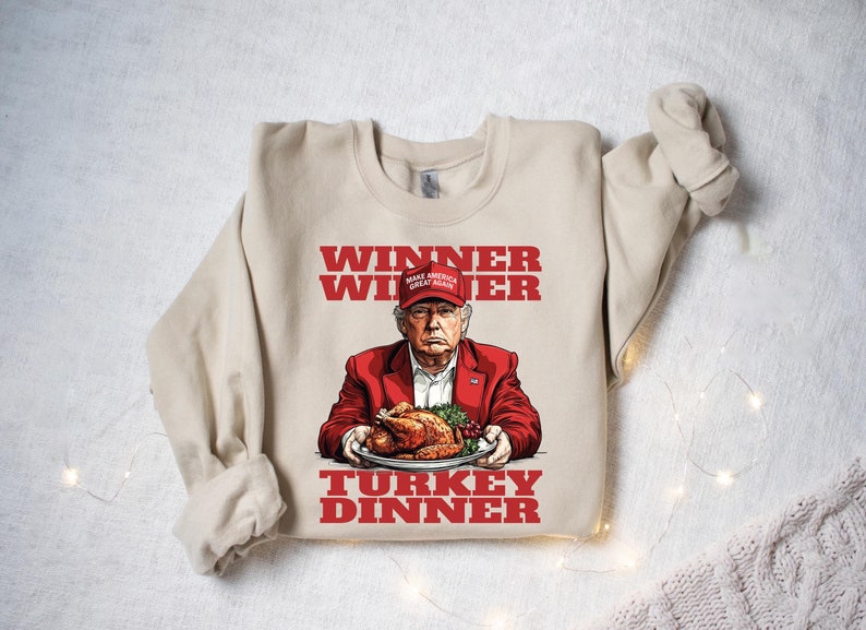 May include: A beige sweatshirt with a red graphic print of a man wearing a red "Make America Great Again" hat holding a plate of roasted turkey. The text on the sweatshirt reads "Winner Winner Turkey Dinner".