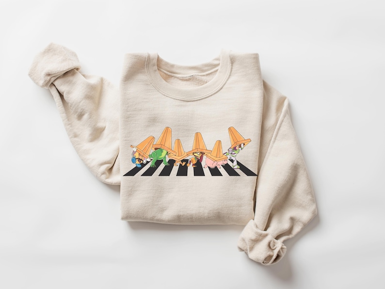 May include: Cream-colored sweatshirt featuring a playful graphic of cartoon characters crossing a black and white striped crosswalk, each wearing an orange traffic cone. The long-sleeved top is folded on a white surface.