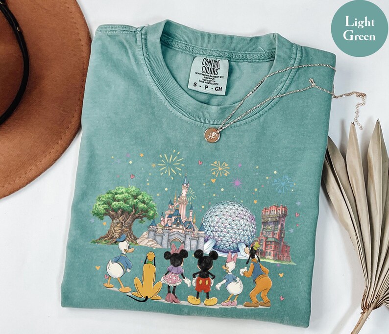 May include: A light green t-shirt featuring a colorful graphic of Disney characters and landmarks, including a castle, Spaceship Earth, and the Tree of Life. The shirt has a crew neck and short sleeves. The shirt is made of a soft material.