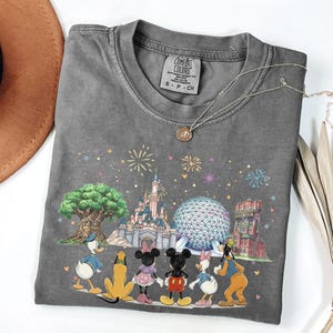 May include: Gray Comfort Colors t-shirt featuring a colorful graphic of Disney characters and landmarks. The design includes Donald Duck, Mickey Mouse, Minnie Mouse, and Goofy, with fireworks and a castle. A silver necklace is draped on the shirt.