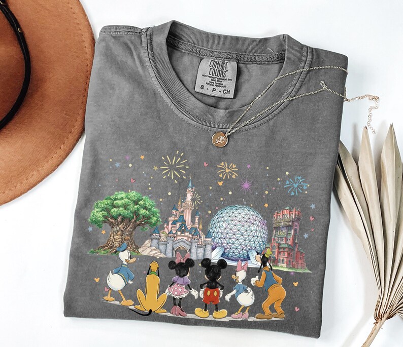 May include: Gray Comfort Colors t-shirt featuring a colorful graphic of Disney characters and landmarks. The design includes Donald Duck, Mickey Mouse, Minnie Mouse, and Goofy, with fireworks and a castle. A silver necklace is draped on the shirt.