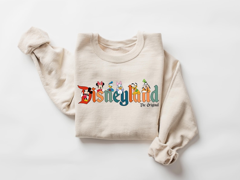 Może przedstawiać: Beżowa bluza z logo Disneyland z Myszka Miki, Myszka Minnie, Kaczorem Donaldem, Daisy, Goofym i Plutonem. Tekst "The Original" znajduje się pod tekstem Disneyland. Bluza jest złożona.