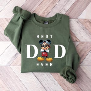 Op de afbeelding: Groen sweatshirt met een witte afbeelding van Mickey Mouse met een zonnebril en de tekst "BEST DAD EVER".