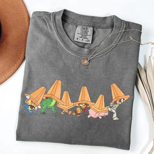 Puede incluir: Una camiseta gris Comfort Colors con un gr&aacute;fico de personajes de dibujos animados debajo de conos de tr&aacute;fico naranjas. La camiseta tiene cuello redondo y una peque&ntilde;a etiqueta en la parte superior de la espalda. Un collar plateado con un colgante est&aacute; sobre la camiseta.