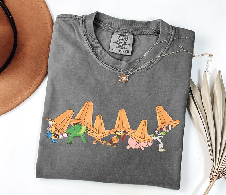 Puede incluir: Una camiseta gris Comfort Colors con un gr&aacute;fico de personajes de dibujos animados debajo de conos de tr&aacute;fico naranjas. La camiseta tiene cuello redondo y una peque&ntilde;a etiqueta en la parte superior de la espalda. Un collar plateado con un colgante est&aacute; sobre la camiseta.