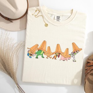 Puede incluir: Camiseta de color crema con un gr&aacute;fico de dibujos animados de personajes que llevan conos de tr&aacute;fico naranjas. Tambi&eacute;n se ven un collar dorado, un sombrero beige con una banda marr&oacute;n y sandalias marrones. La camiseta tiene una etiqueta que dice "Comfort Colors".
