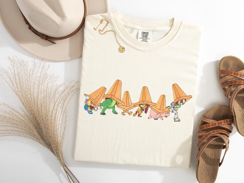 Puede incluir: Camiseta de color crema con un gr&aacute;fico de dibujos animados de personajes que llevan conos de tr&aacute;fico naranjas. Tambi&eacute;n se ven un collar dorado, un sombrero beige con una banda marr&oacute;n y sandalias marrones. La camiseta tiene una etiqueta que dice "Comfort Colors".