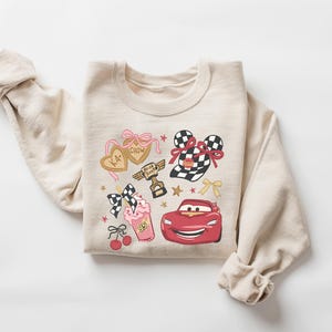 May include: Beige sweatshirt featuring a colorful graphic design. The design includes a red race car, a milkshake, and heart-shaped cookies with text. Additional elements include bows, stars, and a trophy. The sweatshirt has long sleeves.