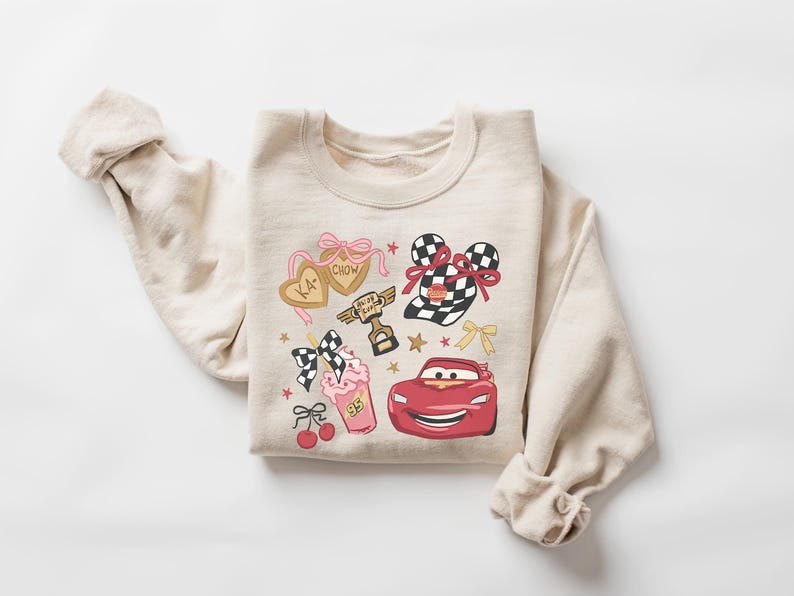 May include: Beige sweatshirt featuring a colorful graphic design. The design includes a red race car, a milkshake, and heart-shaped cookies with text. Additional elements include bows, stars, and a trophy. The sweatshirt has long sleeves.