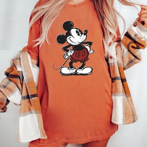 May include: A woman wearing a burnt orange t-shirt with a vintage Mickey Mouse graphic. She is also wearing a plaid flannel shirt.