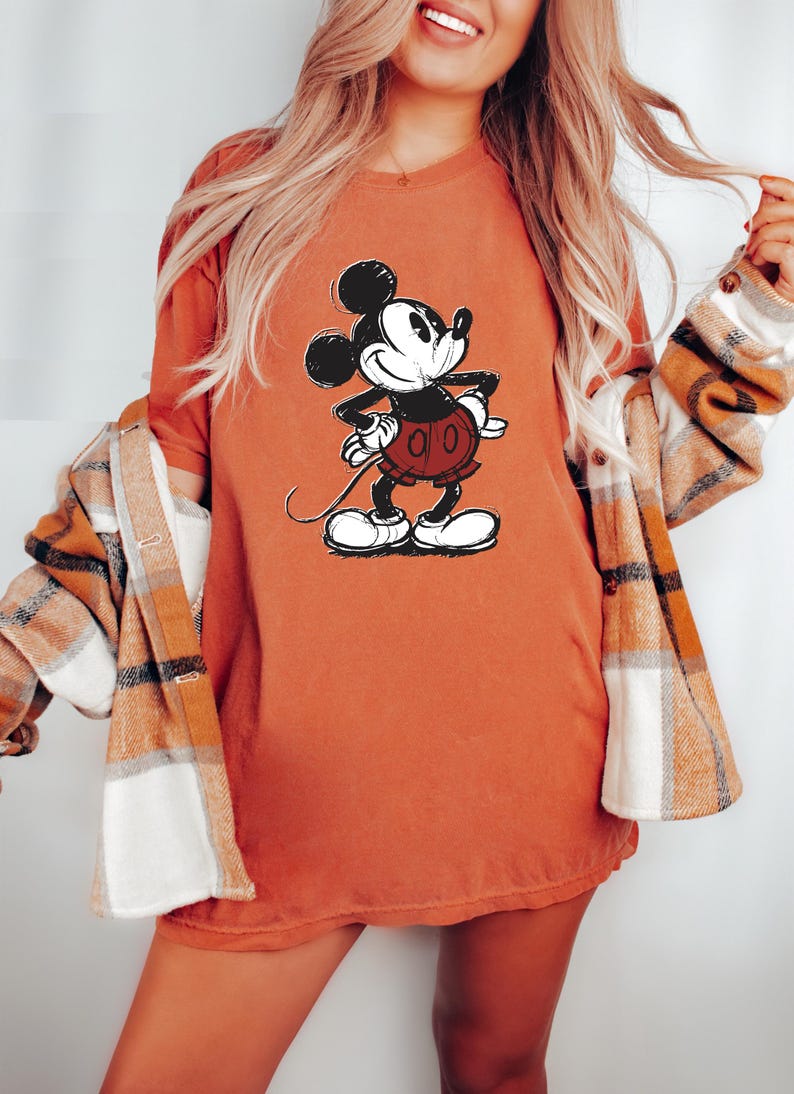 May include: A woman wearing a burnt orange t-shirt with a vintage Mickey Mouse graphic. She is also wearing a plaid flannel shirt.