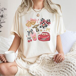 May include: Cream-colored t-shirt featuring a colorful graphic design. The design includes a red race car, a trophy, hearts, bows, and a milkshake. The shirt is a casual, comfortable style.