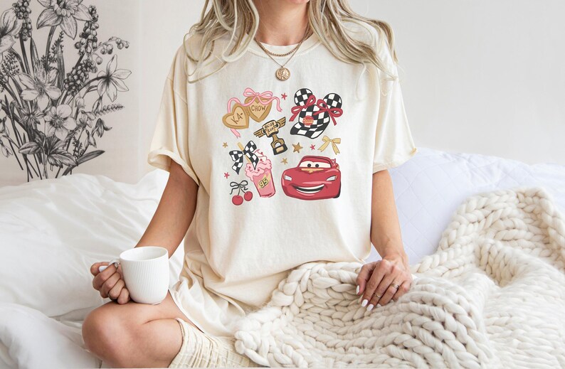 May include: Cream-colored t-shirt featuring a colorful graphic design. The design includes a red race car, a trophy, hearts, bows, and a milkshake. The shirt is a casual, comfortable style.