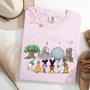 May include: A light pink t-shirt featuring a colorful graphic of Disney characters in front of a castle, tree, and other landmarks. The shirt is paired with brown sandals, a beige hat, and a gold necklace.