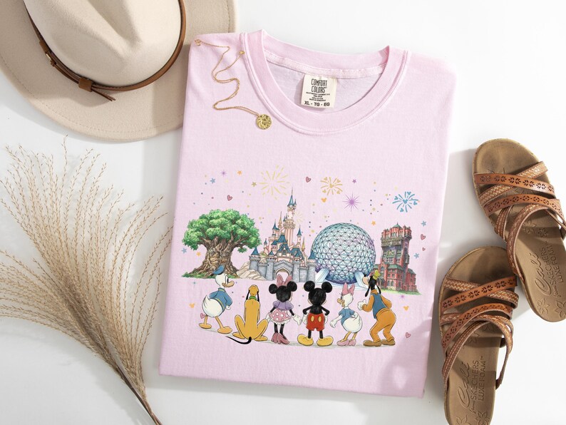 May include: A light pink t-shirt featuring a colorful graphic of Disney characters in front of a castle, tree, and other landmarks. The shirt is paired with brown sandals, a beige hat, and a gold necklace.