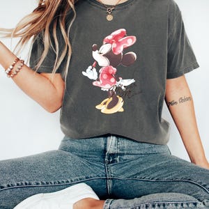 Puede incluir: Una camiseta gris oscuro con un gr&aacute;fico de Minnie Mouse con un vestido rojo de lunares y un lazo rojo. El texto "Minnie Mouse" est&aacute; escrito debajo del gr&aacute;fico.